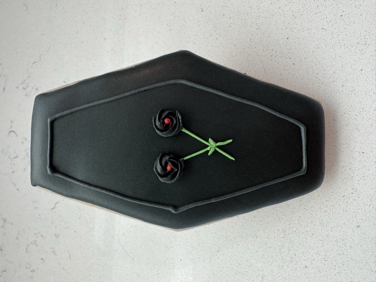 A black coffin-shaped decorated sugar cookie with a small rose and crossed branches design