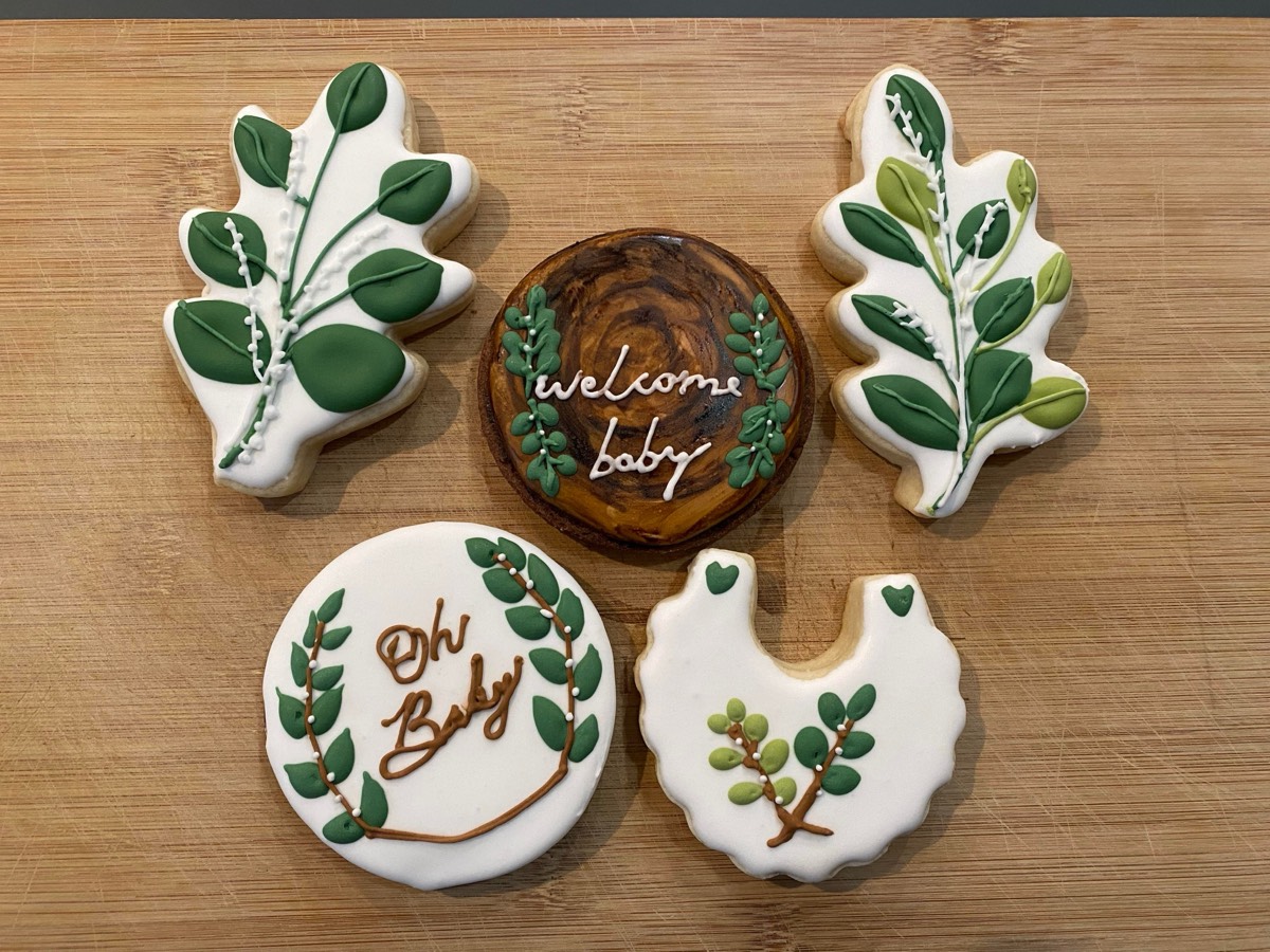 Green botanical baby shower decorated sugar cookies with oak leaf branches, a "Welcome Baby" cookie, an "Oh Baby" wreath cookie, and a onesie cookie