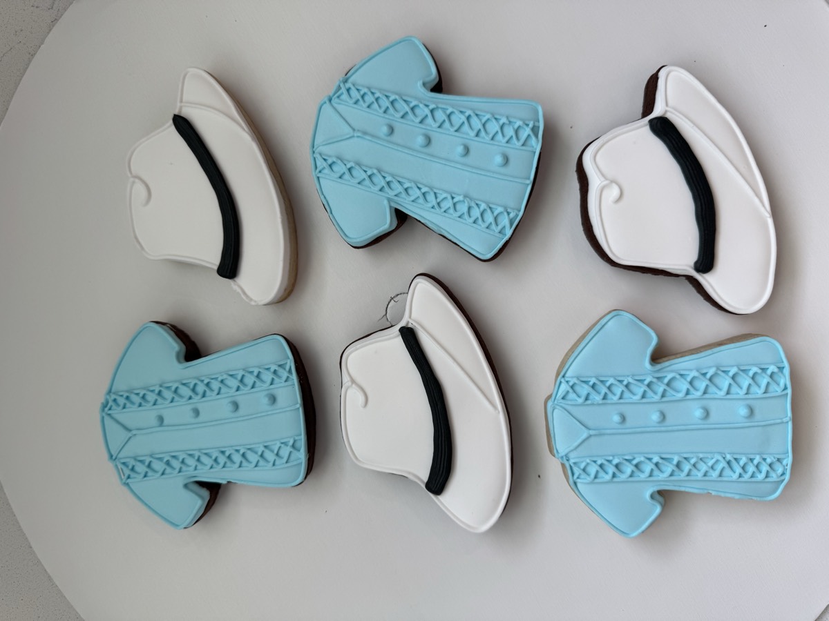 Dapper-themed baby shower sugar cookies shaped like light blue button-down shirts and white fedora hats with black bands