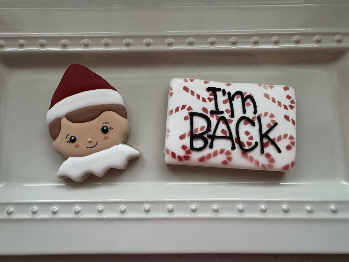 An Elf on the Shelf face decorated sugar cookie next to a candy-cane patterned cookie reading "I'm BACK"