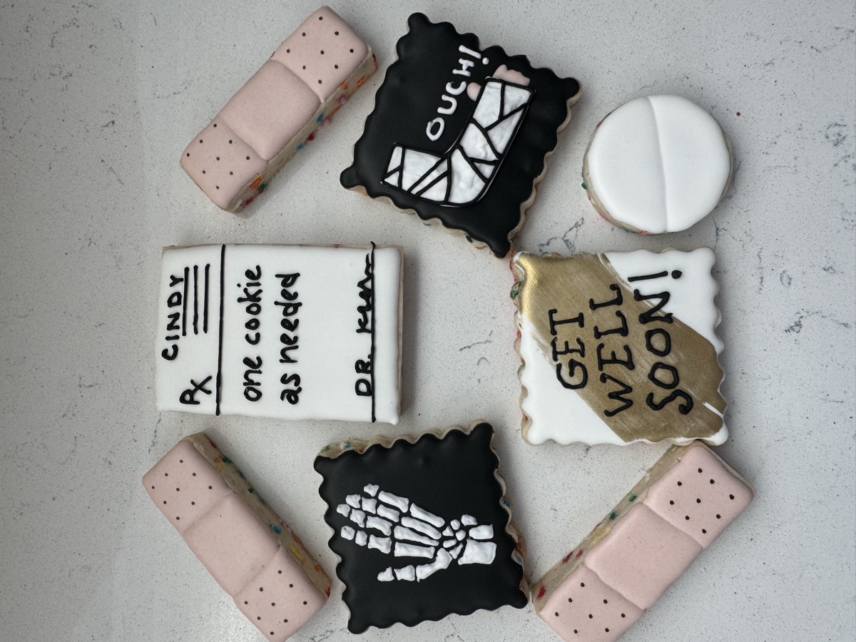 Get well themed decorated sugar cookies: bandages, a prescription pad reading "one cookie as needed", a skeleton hand, a broken-bone cast reading "OUCH!", a gold "GET WELL SOON!" cookie, and a pill