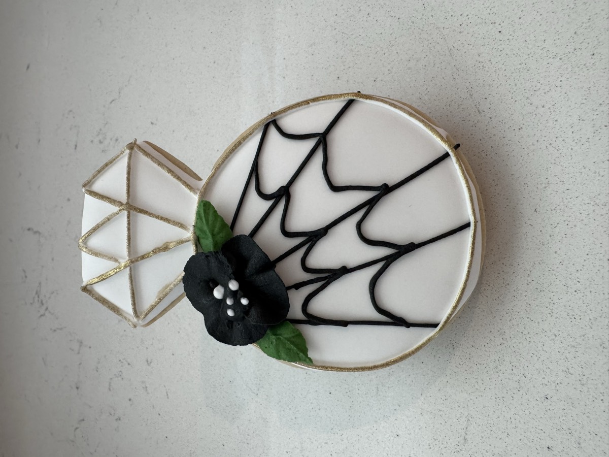 A diamond engagement ring decorated sugar cookie with a black spiderweb and flower design, a Halloween-wedding mashup