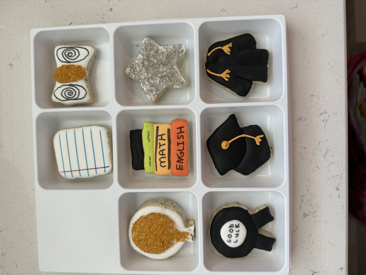 A boxed graduation cookie set with a notebook-paper design, a bow tie, a gold balloon, stacked Math and English books, a silver star, a "Good Luck" medal, a graduation cap, and a cap-and-gown cookie