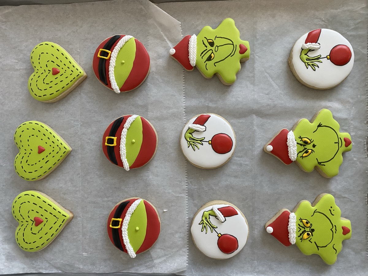 Grinch-themed Christmas decorated sugar cookies featuring the Grinch's face, Santa hat trees, striped hearts, and his hand holding an ornament