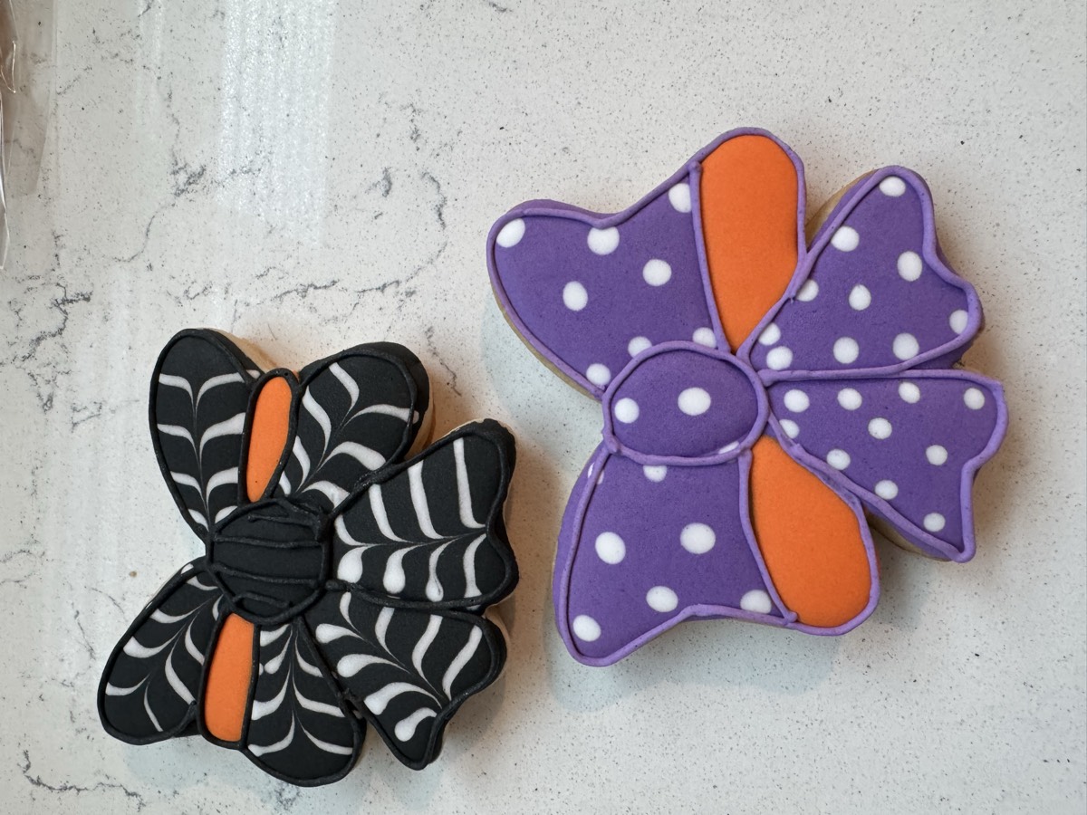 Two Halloween decorated sugar cookie bows, one black-and-white spiderweb patterned and one purple with white polka dots, both with orange centers