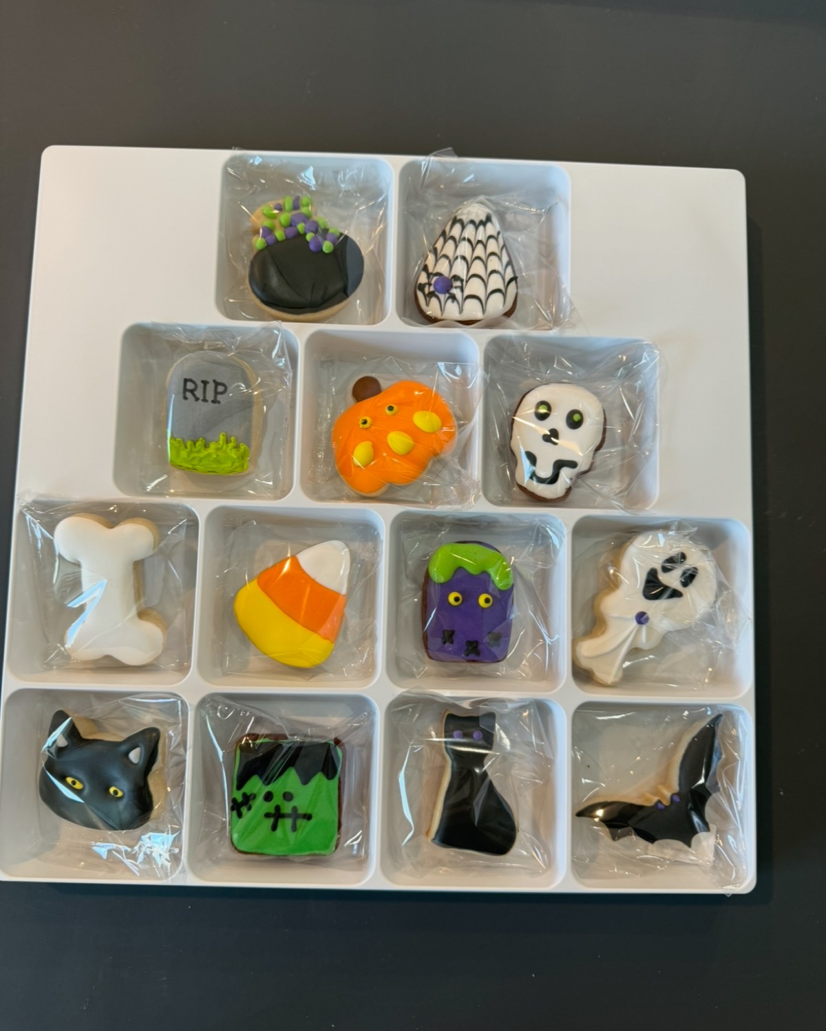 A 12-piece boxed Halloween decorated sugar cookie set with a cauldron, spiderweb, tombstone, pumpkin, skull, bone, candy corn, Frankenstein, black cat, and bat designs