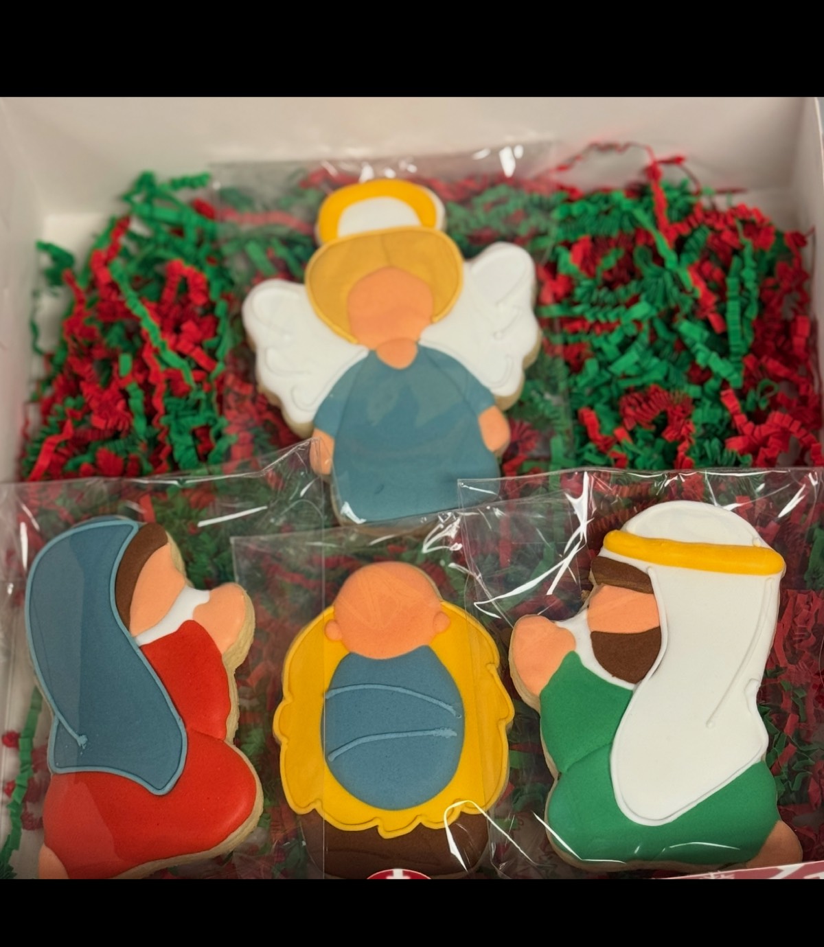 A gift box of nativity decorated sugar cookies with an angel, Mary, baby Jesus, and Joseph, nestled in red and green paper shred