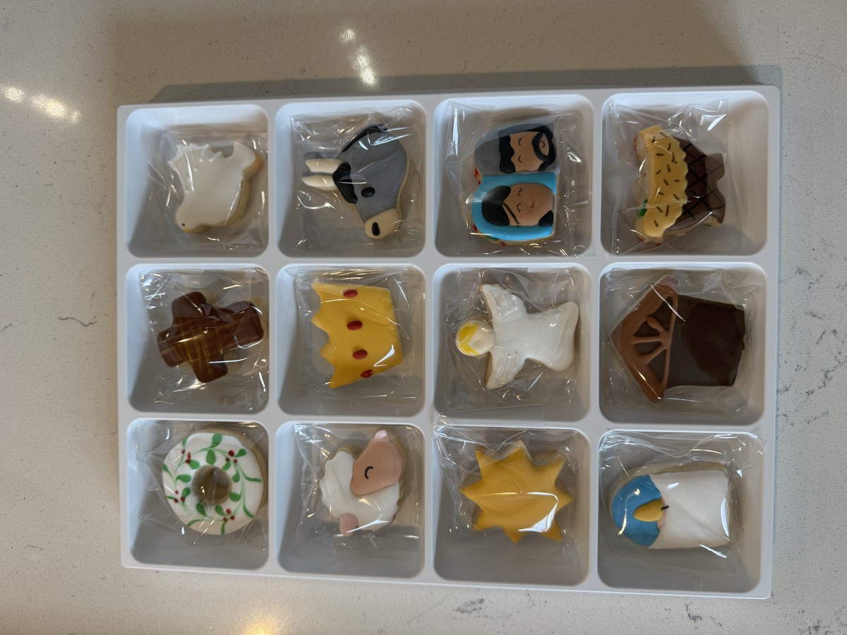 A 12-piece boxed nativity-themed cookie set including a wreath, cross, dove, lamb, crown, star, angel, and manger scene figures