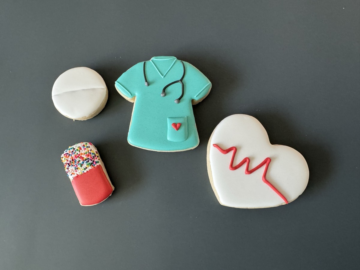 Medical themed decorated sugar cookies: a sprinkle-topped capsule pill, a round pill, a teal nurse scrub top, and an EKG heartbeat heart