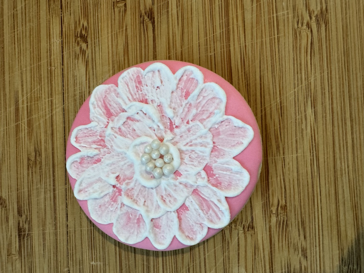 A pink round decorated sugar cookie with white ruffled petals and a pearl cluster center