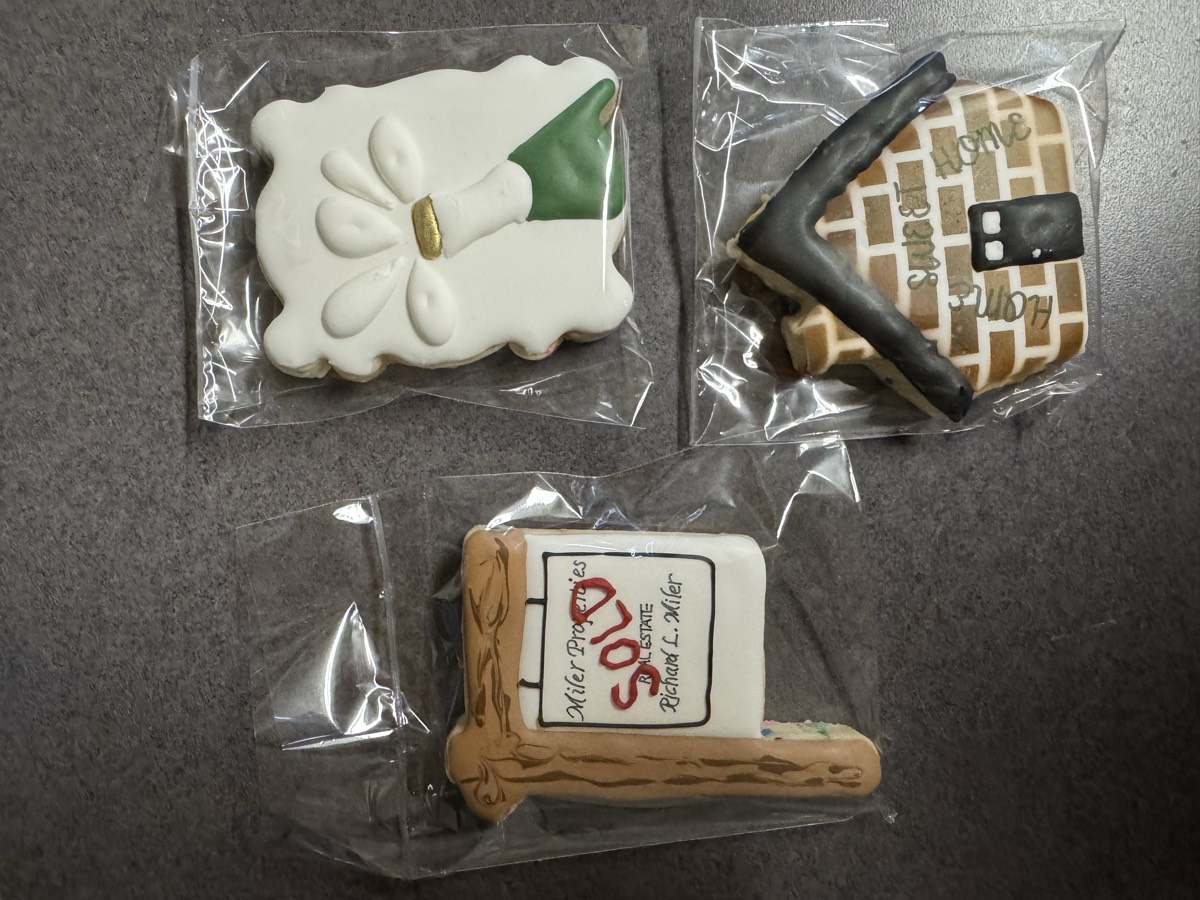 Real estate themed decorated sugar cookies: a "SOLD" yard sign with a realtor's name, a bow-tied gift cookie, and a brick house cookie