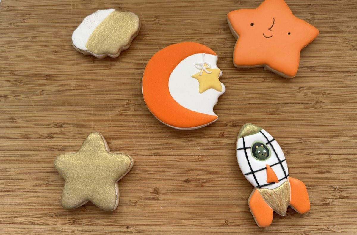 Celestial baby shower decorated sugar cookies: a cloud, a smiling star, a crescent moon with a dangling star, another gold star, and a rocket ship