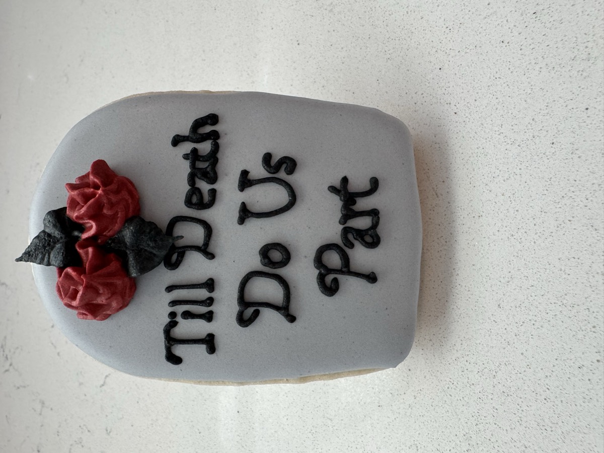 A gray tombstone decorated sugar cookie with red roses reading "Till Death Do Us Part", a Halloween-wedding mashup design