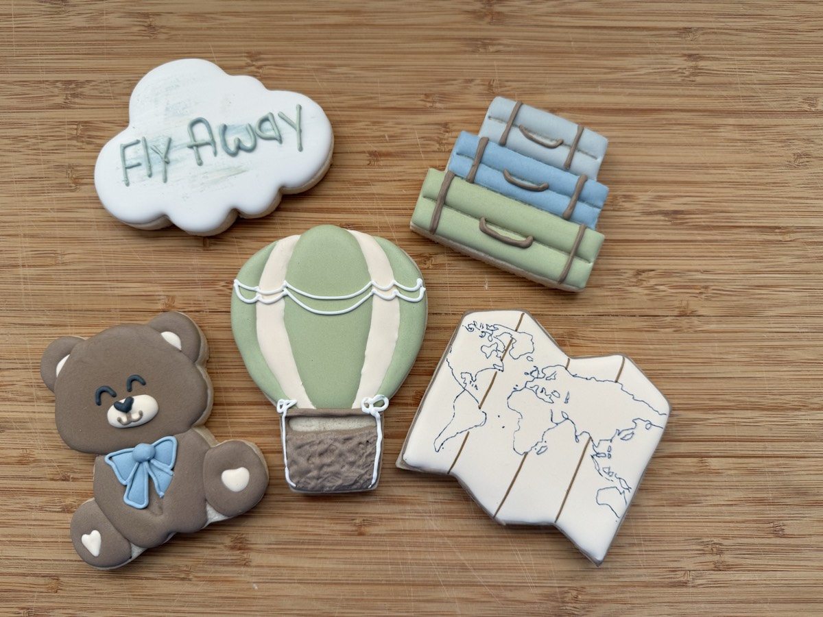Travel themed baby shower decorated sugar cookies: a teddy bear with a blue bow, a "Fly Away" cloud, a hot air balloon, a world map suitcase, and striped travel books