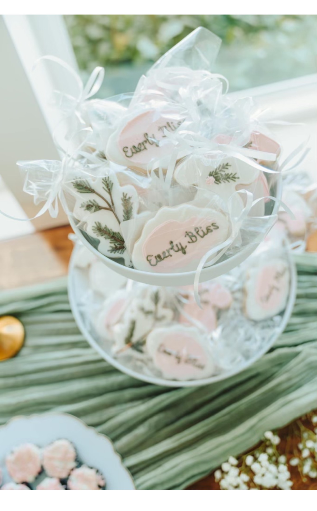 Pink and white "Every Bliss" wedding favor decorated sugar cookies with greenery, displayed on a tiered stand